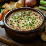 Caramelized onion and garlic dip garnished with chives, served in a ceramic bowl with crackers and fresh vegetables on a wooden board.