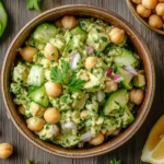 A vibrant bowl of avocado chickpea tuna salad with fresh herbs, lemon slices, and crunchy vegetables.