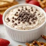 A bowl of creamy cookie dough dip with chocolate chips, surrounded by graham crackers and strawberries.