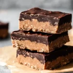 A stack of homemade chocolate peanut butter protein bars on a wooden board, topped with melted dark chocolate and sprinkled with sea salt.