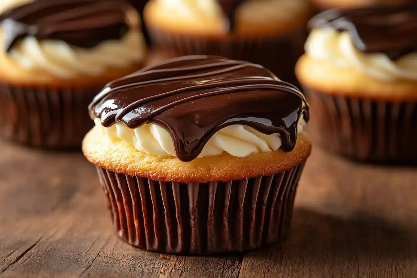 A plate of freshly made Boston Cream Pie Cupcakes with glossy chocolate ganache and creamy filling, set on a rustic wooden table.
