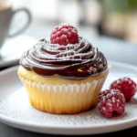 A Boston Cream Pie Cupcake on a white plate, dusted with powdered sugar, garnished with a fresh raspberry, and served with a hot espresso.
